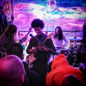 guitarists and drummer performing in front of projector screen and audience