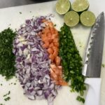 cutting board with cilantro, red onions, tomatoes, and jalapeño diced, two limes cut in half and a large knife